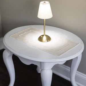 May include: A white oval side table with curved legs, topped with a cream-colored rectangular placemat. A small brass lamp with a pleated shade sits on the placemat, casting a warm glow. The table is set against a neutral wall and dark wood floor.