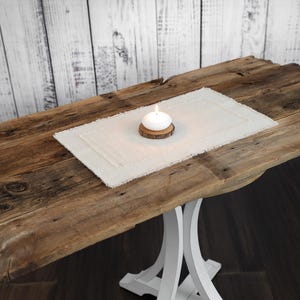 May include: A rustic wooden table with a white base. A white linen placemat with a fringed edge sits on the table, holding a lit white candle on a wooden coaster. The background is a white wooden wall.