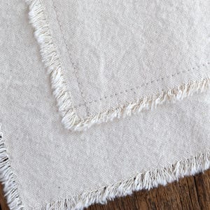 Puede incluir: Primer plano de dos servilletas de lino blanquecino con bordes deshilachados. Las servilletas est&aacute;n hechas de una tela texturizada y tienen una est&eacute;tica sencilla y r&uacute;stica. Los bordes est&aacute;n deshilachados, a&ntilde;adiendo un toque de elegancia informal.