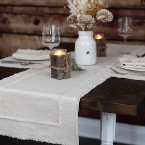 May include: A dining table set for a meal. The table is covered with a cream-colored runner and placemats. A vase of dried flowers, candles, and glassware are arranged on the table. The table is dark brown with white legs.