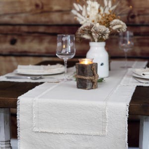 May include: A rustic dining table setting with a cream-colored linen table runner. A lit candle in a wooden holder sits on the runner, along with a white vase of dried flowers and two wine glasses. The table is set for a meal.