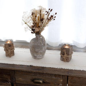 May include: A rustic home decor arrangement featuring a weathered gray vase with dried flowers, flanked by two wooden candle holders wrapped with rope. The items are arranged on a cream-colored table runner atop a wooden dresser.