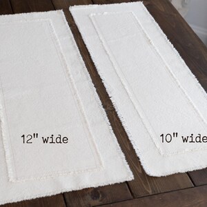 Puede incluir: Dos caminos de mesa rectangulares de color blanquecino con borde deshilachado se muestran sobre una superficie de madera oscura. Uno est&aacute; etiquetado como "12" wide" y el otro como "10" wide". El m&aacute;s ancho mide unos 30 cm de ancho.
