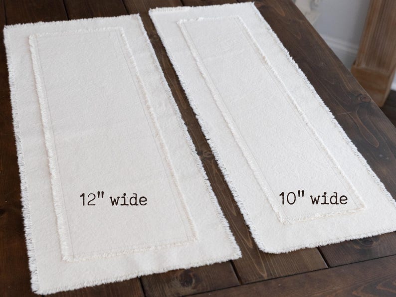 Puede incluir: Dos caminos de mesa rectangulares de color blanquecino con borde deshilachado se muestran sobre una superficie de madera oscura. Uno est&aacute; etiquetado como "12" wide" y el otro como "10" wide". El m&aacute;s ancho mide unos 30 cm de ancho.