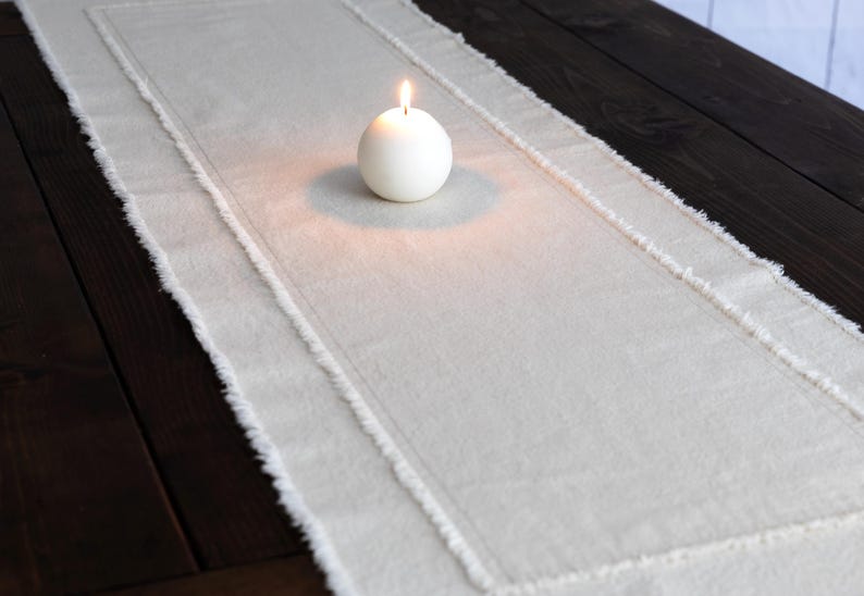Puede incluir: Un camino de mesa color crema con borde deshilachado y borde cosido. Una vela blanca esf&eacute;rica con una llama encendida se encuentra en el centro del camino de mesa. El camino de mesa est&aacute; colocado sobre una mesa de madera oscura.