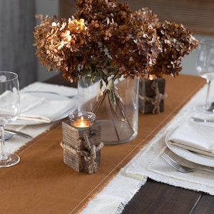 May include: A dining table setting with a brown table runner, white placemats, and clear wine glasses with white polka dots. A vase of dried flowers and wooden candle holders with rope accents are also present.