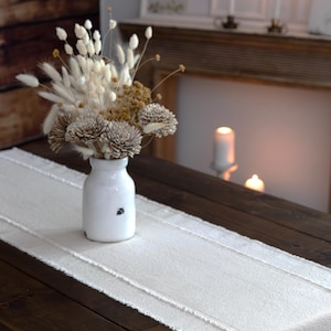 May include: A white ceramic vase with a bee design holds a dried flower arrangement on a cream-colored table runner. The runner sits on a dark brown wooden table. Candles and a decorative frame are in the background.