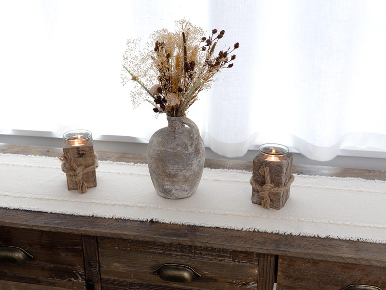 Puede incluir: Una composici&oacute;n decorativa r&uacute;stica con un jarr&oacute;n gris envejecido lleno de flores secas, flanqueado por dos candelabros de madera atados con cuerda. Las velas encendidas crean un ambiente c&aacute;lido. El conjunto est&aacute; sobre un camino de mesa crema.