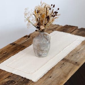 May include: A rustic wooden table with a cream-colored linen table runner and a gray ceramic vase holding dried flowers. The table runner has a fringed edge, and the vase has a handle.