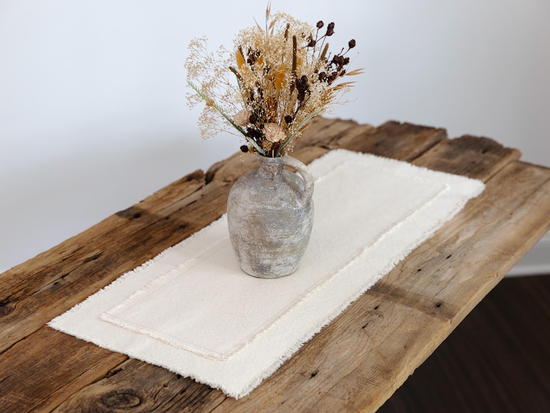 Puede incluir: Una mesa de madera r&uacute;stica con un camino de mesa de lino color crema y un jarr&oacute;n de cer&aacute;mica gris con flores secas. El camino de mesa tiene un borde con flecos y el jarr&oacute;n tiene un asa.