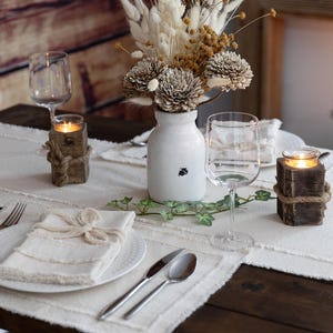 Puede incluir: Una mesa r&uacute;stica con un camino de mesa blanco, platos y servilletas. Incluye copas de vino, cubiertos, un jarr&oacute;n blanco con flores secas y candelabros de madera atados con cuerda. La luz de las velas crea un ambiente c&aacute;lido.