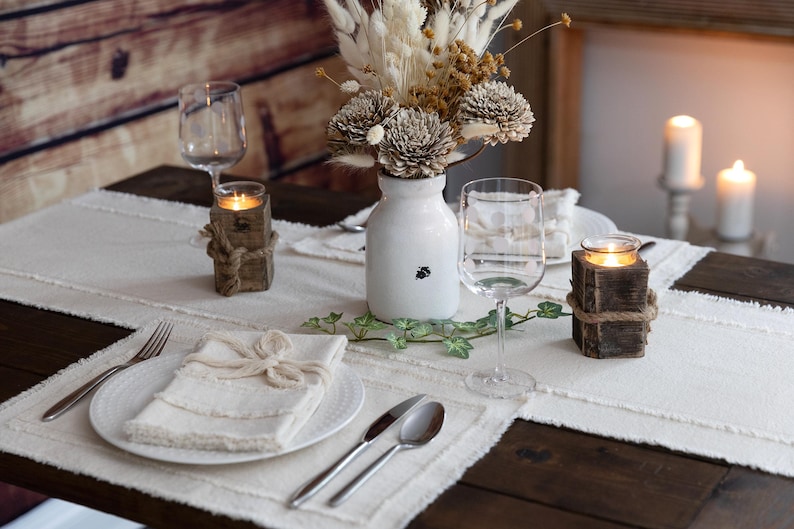Puede incluir: Una mesa r&uacute;stica con un camino de mesa blanco, platos y servilletas. Incluye copas de vino, cubiertos, un jarr&oacute;n blanco con flores secas y candelabros de madera atados con cuerda. La luz de las velas crea un ambiente c&aacute;lido.