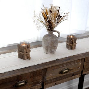 May include: A rustic wooden console table with a cream-colored runner and decorative items. The table features a distressed finish and brass drawer pulls. Two wooden candle holders wrapped with rope flank a vase filled with dried flowers.