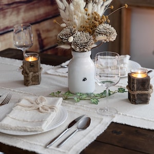 May include: A rustic dining table setting with a white runner, plates, and napkins. Includes wine glasses, silverware, a white vase with dried flowers, and wooden candle holders wrapped with rope. Soft lighting from candles creates a warm ambiance.