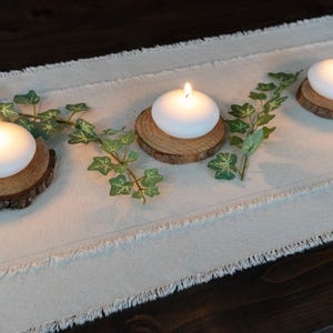 Puede incluir: Tres velas blancas y redondas con llamas encendidas se asientan sobre peque&ntilde;as rodajas de madera r&uacute;stica. Acentos de hiedra verde se colocan entre las velas sobre un camino de mesa de lino color crema. El arreglo est&aacute; sobre una superficie de madera oscura.