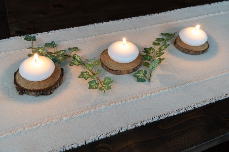 Puede incluir: Tres velas blancas y redondas con llamas encendidas se asientan sobre peque&ntilde;as rodajas de madera r&uacute;stica. Acentos de hiedra verde se colocan entre las velas sobre un camino de mesa de lino color crema. El arreglo est&aacute; sobre una superficie de madera oscura.