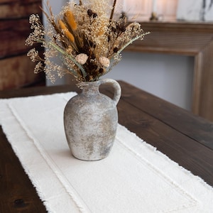 May include: A rustic, gray ceramic vase with a handle holds a bouquet of dried flowers in shades of beige, brown, and yellow. The vase sits on a white linen table runner with a fringed edge, placed on a dark wooden table.