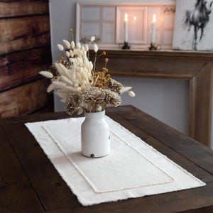 May include: A white ceramic vase holds a dried floral arrangement on a cream-colored table runner. The runner sits on a dark wooden table. The arrangement includes beige, brown, and tan dried flowers. The background features a wooden mantel and artwork.