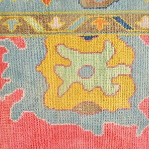 May include: Close-up of a colorful rug with a detailed pattern. The rug features a pink field with light blue and yellow geometric shapes. A border with brown, yellow, and blue accents frames the design. The rug has a soft, textured appearance.