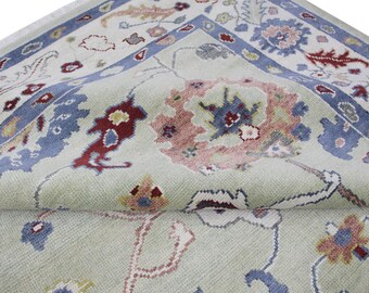 May include: Close-up of a rolled-up area rug with a floral pattern. The rug features a light green background with a blue border and accents in red, pink, and yellow. The design includes stylized floral and leaf motifs.