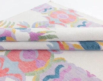 May include: Folded white fabric with a colorful floral pattern. The design features pink, purple, blue, and yellow elements. The fabric is layered, showing the pattern on the top and edges.