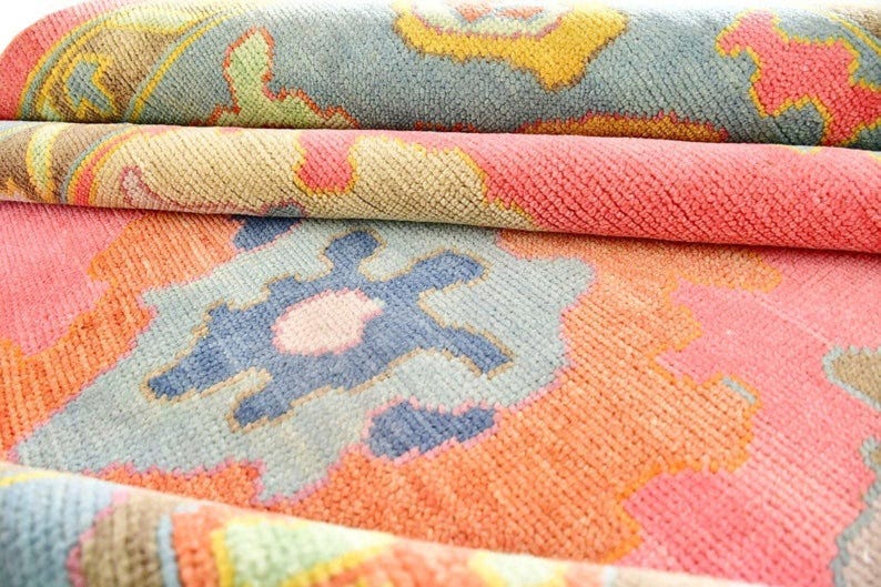 May include: Close-up of a rolled-up rug with a colorful, intricate pattern. The rug features a mix of coral, blue, yellow, and green hues. The design includes floral and geometric shapes, creating a textured and layered appearance.