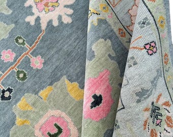 May include: A folded, blue-grey rug with a floral pattern. The rug features large, stylized flowers in pink, yellow, and white, with green and black accents. The border has a repeating floral design. The rug is made of a soft, woven material.