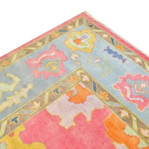 May include: Close-up of a corner of a colorful rug with a light blue border. The rug features a pink and orange center with floral and geometric patterns in pink, yellow, and blue. The border is a light blue with a brown trim.