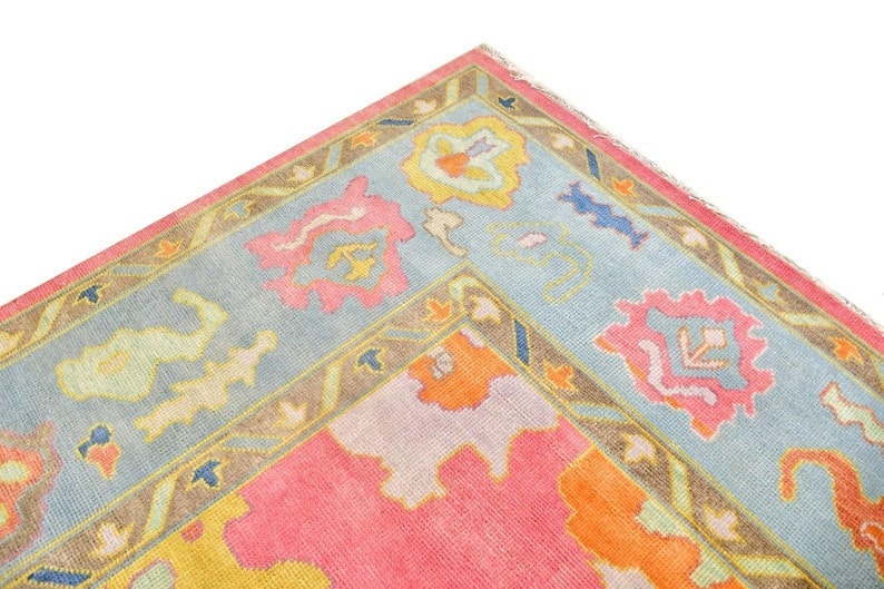 May include: Close-up of a corner of a colorful rug with a light blue border. The rug features a pink and orange center with floral and geometric patterns in pink, yellow, and blue. The border is a light blue with a brown trim.