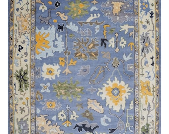 May include: A rectangular area rug with a light blue field and a cream-colored border. The rug features a repeating pattern of floral and abstract designs in yellow, green, and gray. The rug has a fringed edge.