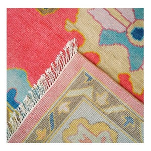 May include: A vibrant, hand-knotted rug with a coral-pink field and intricate floral patterns in shades of blue, yellow, and pink. The rug's border is a soft pink, and it has white fringe.