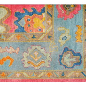 May include: Close-up of a colorful rug with a floral pattern. The rug features a pink border, a light blue center, and various floral designs in shades of pink, blue, yellow, and orange. The rug has a fringed edge.