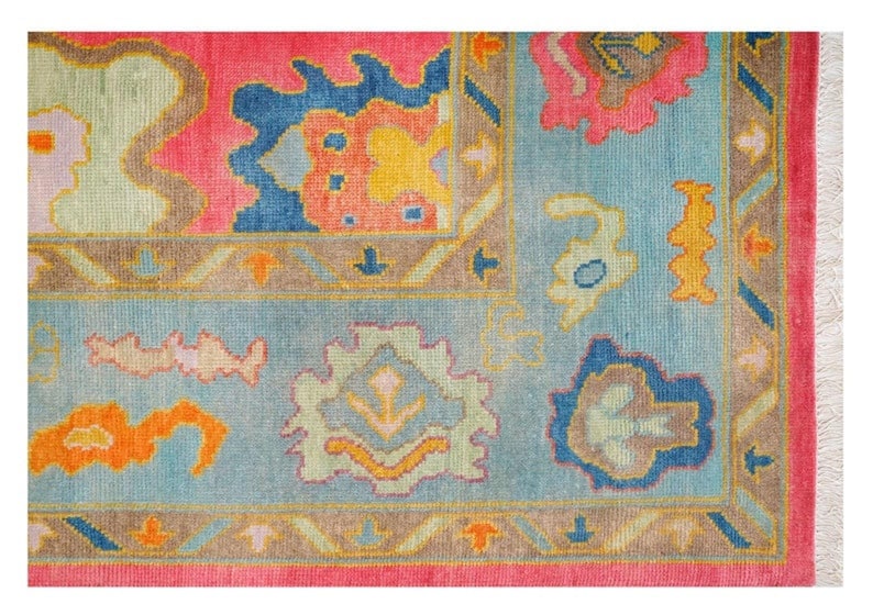 May include: Close-up of a colorful rug with a floral pattern. The rug features a pink border, a light blue center, and various floral designs in shades of pink, blue, yellow, and orange. The rug has a fringed edge.