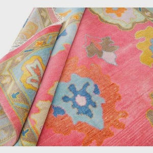 May include: A rolled-up, vibrant pink rug with a detailed floral pattern in shades of blue, yellow, and orange. The rug's border features a complementary design with a light blue and yellow color scheme. The rug is made of a soft material.