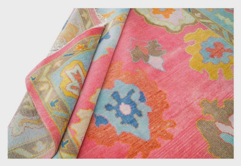 May include: A rolled-up, vibrant pink rug with a detailed floral pattern in shades of blue, yellow, and orange. The rug's border features a complementary design with a light blue and yellow color scheme. The rug is made of a soft material.