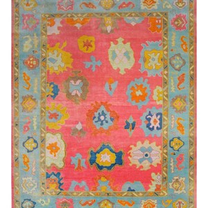 May include: A rectangular rug with a vibrant pink field and a light blue border. The rug features colorful, abstract floral and geometric patterns in shades of yellow, orange, blue, and green. The rug's design is symmetrical, with a central field and a detailed border.
