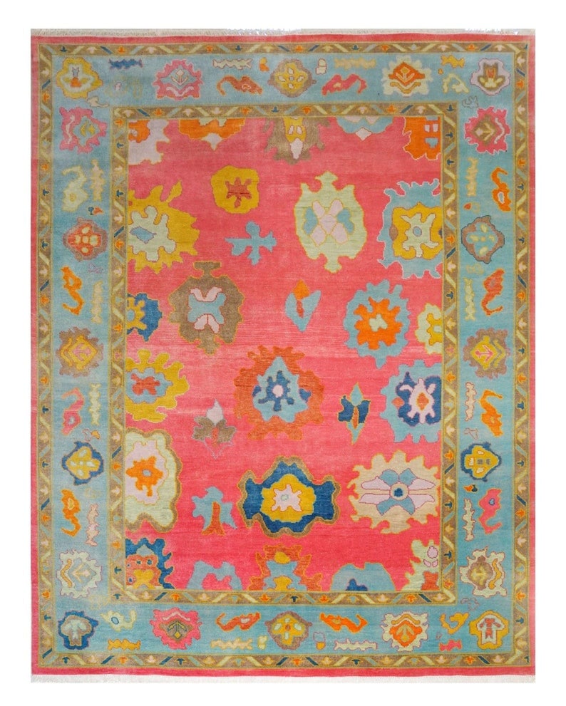 May include: A rectangular rug with a vibrant pink field and a light blue border. The rug features colorful, abstract floral and geometric patterns in shades of yellow, orange, blue, and green. The rug's design is symmetrical, with a central field and a detailed border.