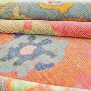 May include: Close-up of a rolled-up area rug with a colorful, patterned design. The rug features a mix of pink, orange, blue, yellow, and green hues. The design includes floral and geometric shapes, creating a vibrant and textured appearance.