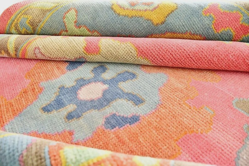 May include: Close-up of a rolled-up area rug with a colorful, patterned design. The rug features a mix of pink, orange, blue, yellow, and green hues. The design includes floral and geometric shapes, creating a vibrant and textured appearance.