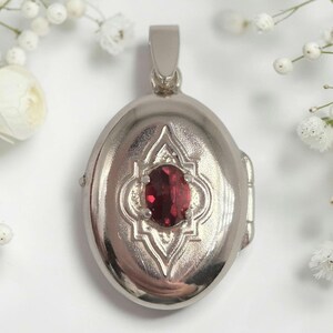 May include: An oval silver-coloured metal locket featuring a central, faceted, deep red oval gemstone set within a decorative, raised geometric frame. The locket has a smooth, polished finish and includes a sturdy bail at the top for suspension.