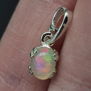 Könnte beinhalten: Ein kleiner ovaler Anhänger mit einem durchscheinenden weißen Opal, der irisierende Blitze in Rosa, Grün und Gelb aufweist. Der Edelstein ist in einer polierten Silberfassung mit Krappen und einer passenden silbernen Öse zur Befestigung gefasst.