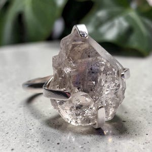 Op de afbeelding: Een zilveren ring met een grote, ruwe, heldere kwartskristal als middelpunt. Het kristal heeft een onregelmatige, veelzijdige vorm met zichtbare interne insluitingen en wordt vastgehouden door vier gepolijste zilveren klauwen op een gladde band.