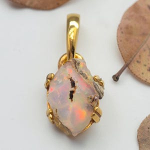 Op de afbeelding: Een hanger van ruwe, doorschijnende opaal in een gepolijste, goudkleurige metalen zetting met klauwen. De opaal vertoont iriserende oranje, groene en roze lichtflitsen op een melkachtige witte basis, met een natuurlijke, onregelmatige vorm.