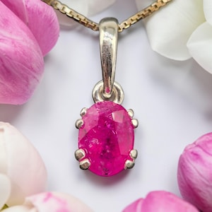 May include: A silver pendant featuring a faceted oval-shaped vibrant pink gemstone held by four prongs. The pendant is attached to a metallic chain link bail, showcasing a polished silver finish and a clear, brilliant cut on the gemstone surface.