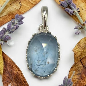 Può includere: Un ciondolo di forma ovale con una pietra sfaccettata e traslucida color azzurro, incastonata in una montatura in metallo argentato smerlato. La pietra presenta inclusioni interne e una superficie lucida, sospesa a una semplice contromaglia liscia.