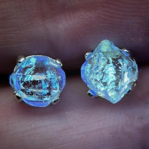 Könnte beinhalten: Ein Paar Ohrstecker mit rohen, durchscheinenden Kristallen, die in einem leuchtenden Blau und irisierend schimmern. Jeder Kristall ist in einer metallischen Krappenfassung befestigt und zeigt eine raue, natürliche Form mit Textur.