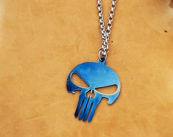 Pode incluir: Um colar de corrente de prata com um pingente de caveira de metal azul. A caveira tem um design simples com duas órbitas e uma boca.