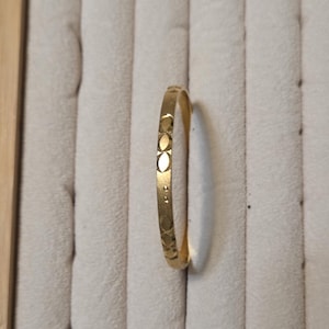 May include: A gold-colored ring with a repeating pattern of small oval shapes. The ring is set against a neutral-toned, textured background, highlighting its simple design and metallic sheen. The ring's surface reflects light, showcasing its polished finish.