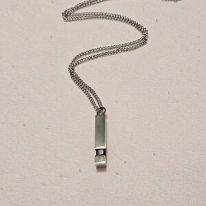 Puede incluir: Un collar de plata con un colgante rectangular. El colgante tiene un pequeño rectángulo negro en el centro.