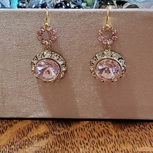 May include: A pair of gold dangle earrings with a large pink crystal surrounded by smaller clear crystals. The earrings have a smaller pink crystal circle above the larger crystal.